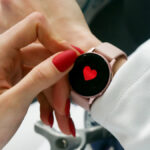 Close-up of a person with red nails touching a smartwatch displaying a red heart on the screen.