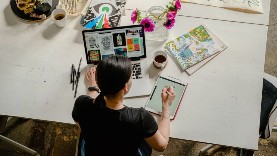 A person is seated at a white table working on a tablet with a stylus, with an open laptop, a notebook, a cup of coffee, and some flowers nearby.