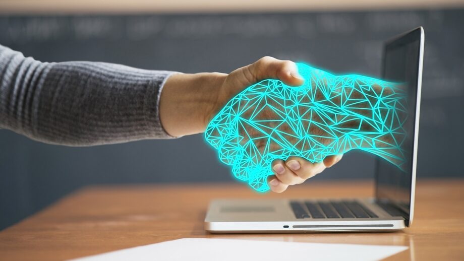 A person's hand extends towards a laptop screen with a graphic of a blue, geometric handshake appearing, symbolizing digital connectivity or online agreements.