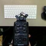 A hand holding a zoom h5 audio recorder with a crossed pair of microphones above a white keyboard and a black computer mouse on a desk.