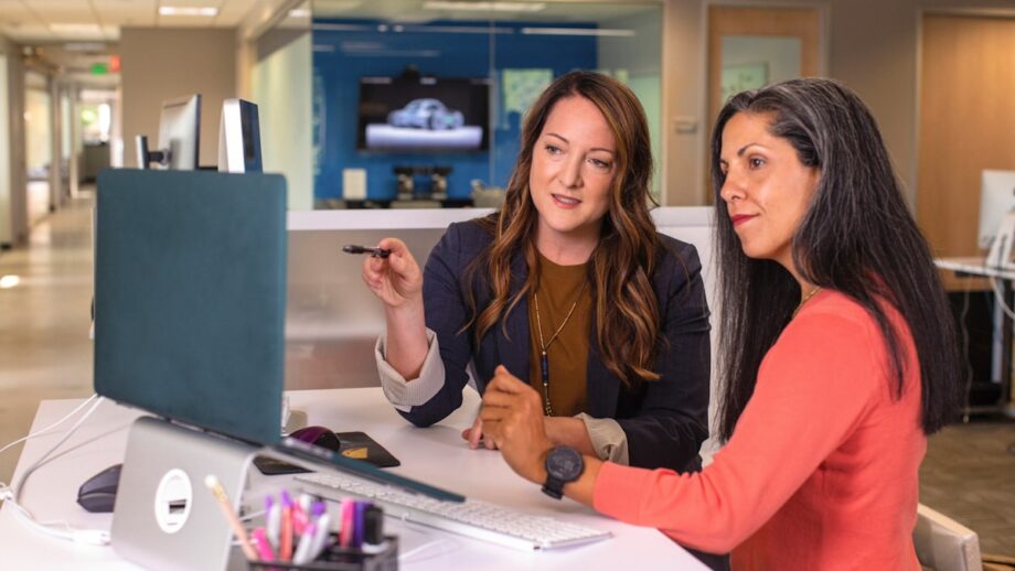 Two women work together at a desk in a bright office, with one pointing at a computer screen and explaining something to the other, who is listening intently.