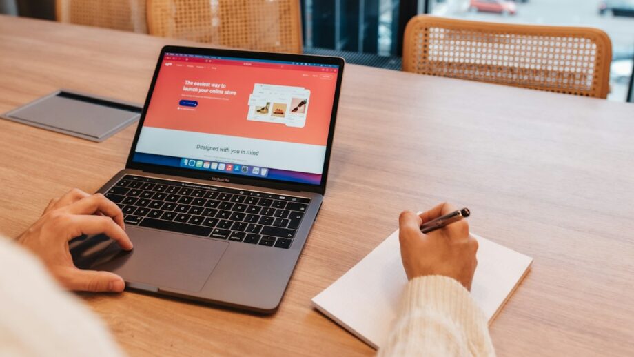 Person using a laptop with an online shop webpage and writing in a notebook, seated at a wooden table in a modern office space with large windows overlooking a body of water.