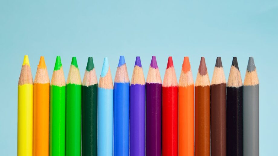 A horizontal row of colored pencils on a light blue background, arranged in a spectrum from yellow to black.