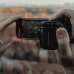 A person holds a digital camera, framing a landscape view of a canyon in the background.