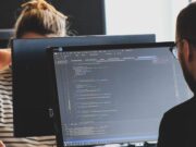 Two people work on computers in an office. The foreground screen displays code in a text editor.