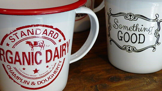 Two vintage-style mugs on a wooden surface. One mug has "Organic Dairy" branding, and the other reads "Something Good.