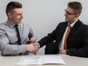 Two men sitting at a table shake hands over a document, wearing formal attire, in a professional setting.