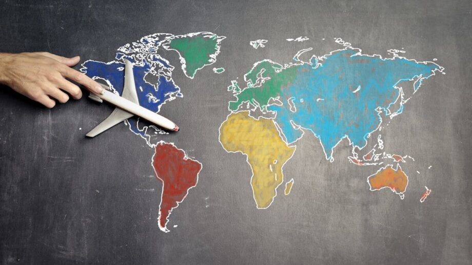 A hand holds a small toy airplane over a chalkboard world map with continents in different colors.