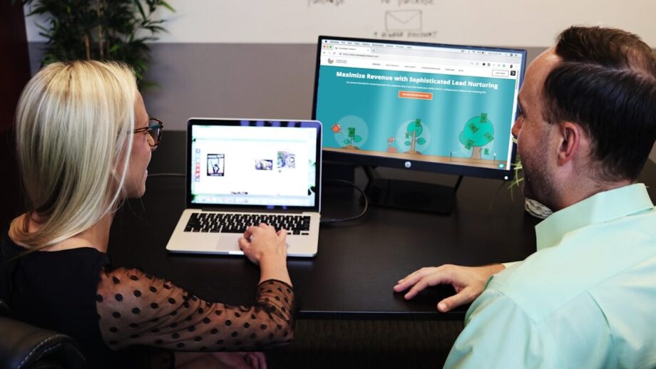 Two people sit at a desk, viewing a laptop and a monitor displaying a lead nurturing website.