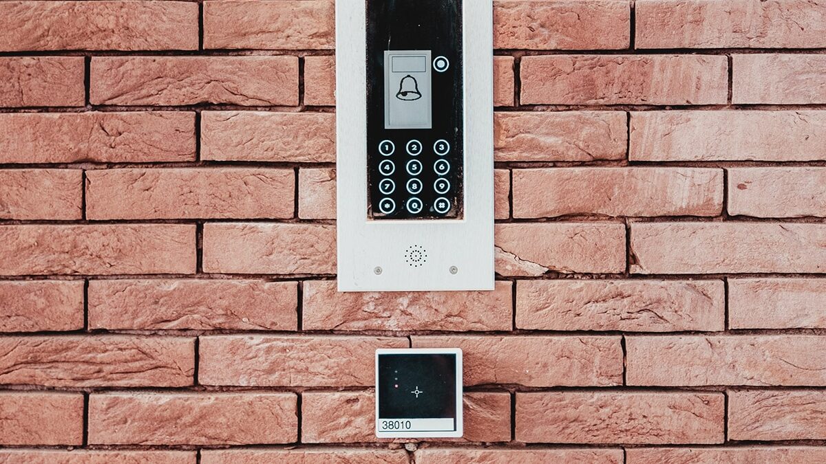 Intercom system mounted on a brick wall, featuring a numeric keypad and bell symbol display, with a small black box below.