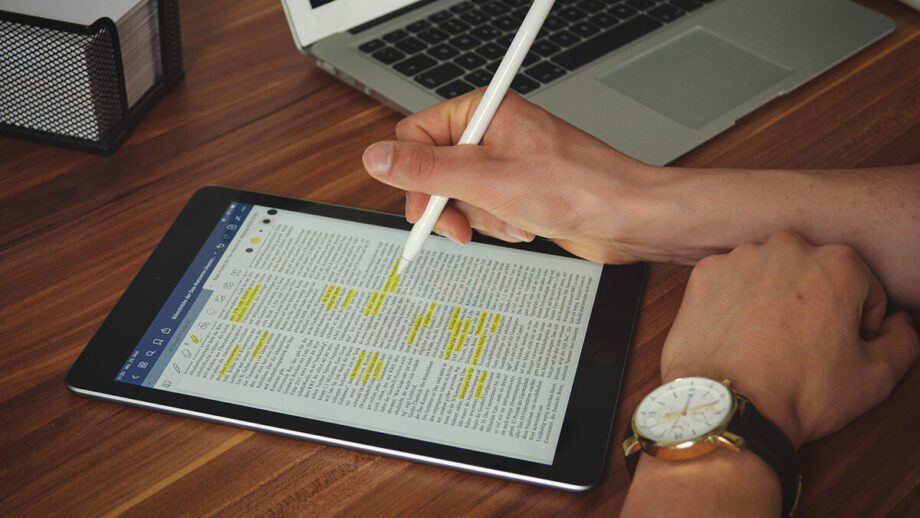 A person uses a stylus on a tablet displaying a document with highlighted text. A laptop and a wire mesh organizer are on the wooden table.