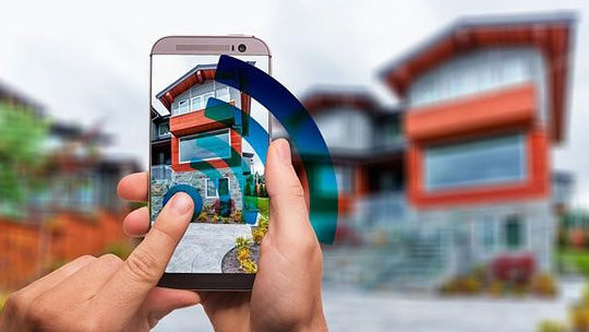 Person holding a smartphone in front of a modern house, capturing it with a Wi-Fi symbol overlay on the screen.