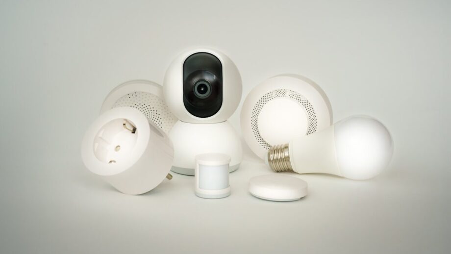 A variety of white smart home devices including a camera, speaker, plug, light bulb, and sensor are displayed against a white background.